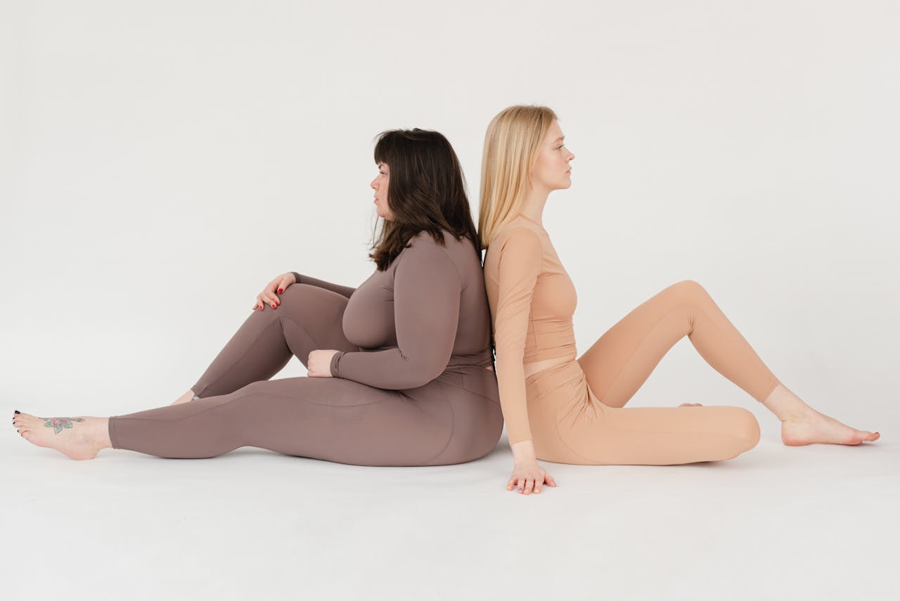 Side view of calm young plus size and fit barefooted female models sitting back to back on floor and looking away against white background